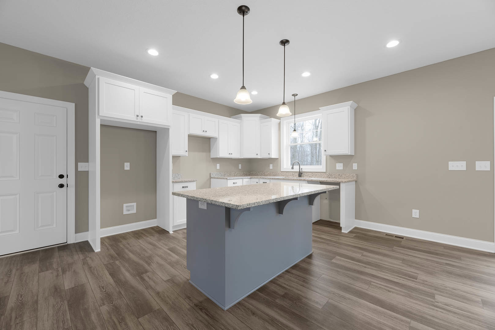Kitchen with white shaker cabinets, large central island featuring granite countertops, stainless steel sink, wood flooring, white door with black hardware, pendant light on black