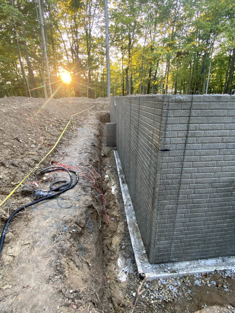 Construction site with exposed concrete foundation, soil, and partially built brick wall, surrounded by trees with sunlight filtering through branches in the background
