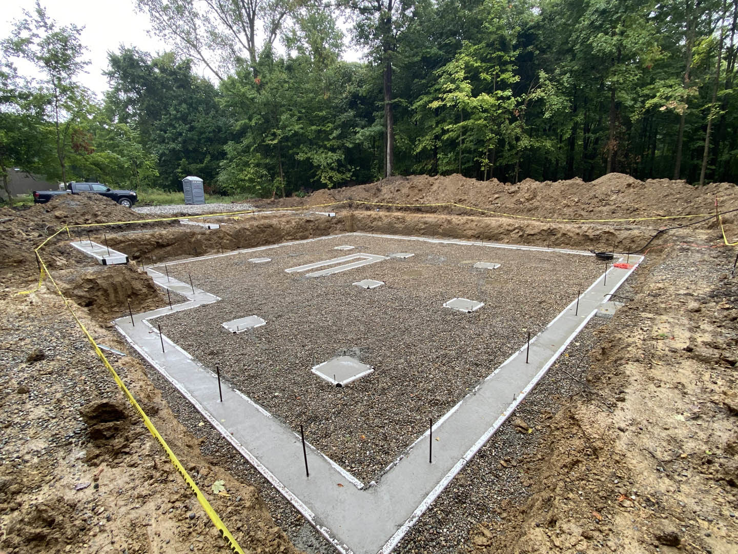 Square concrete foundation surrounded by gravel and soil, portable toilet and parked car nearby, dirt road and trees in background
