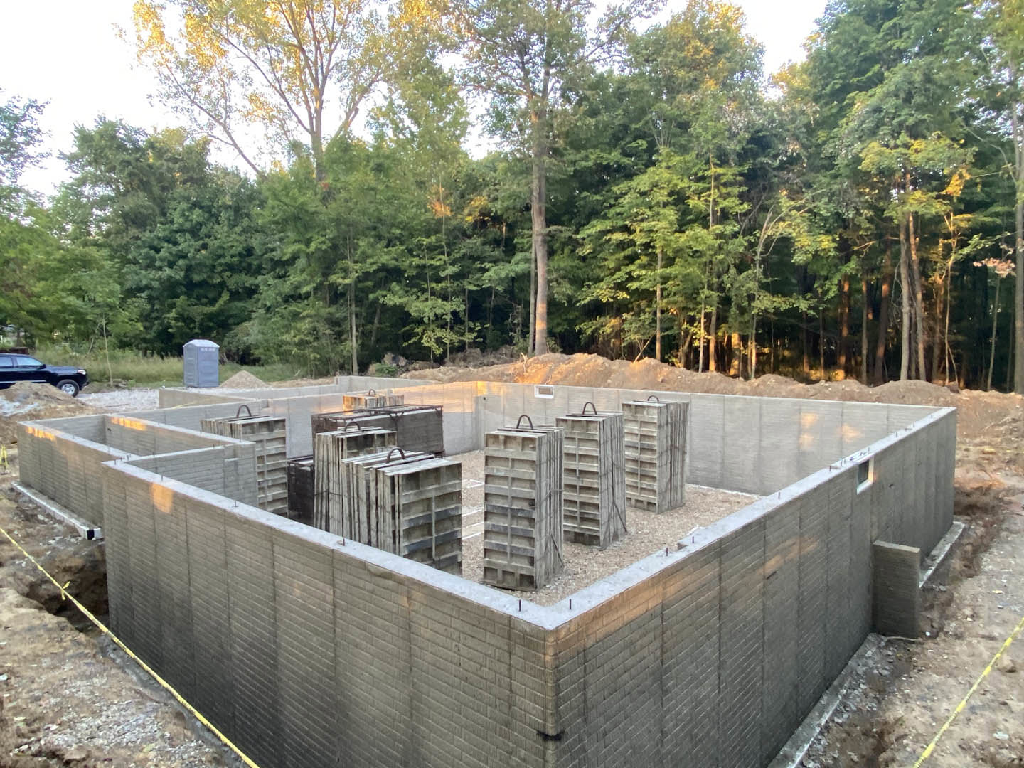 Concrete foundation with exposed wood beams at center, surrounded by dirt ground, portable toilet, parked black car, and trees in background.