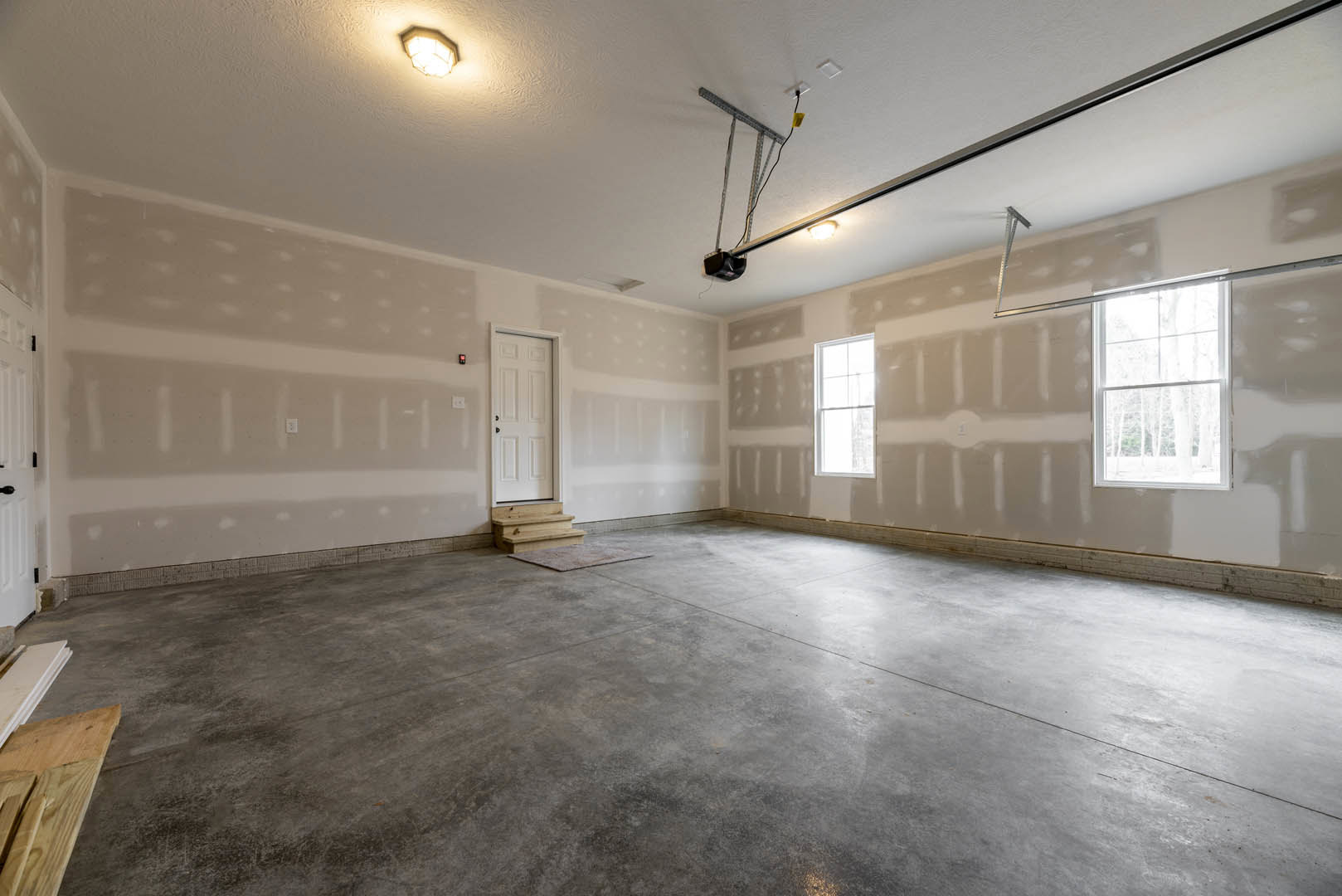 White-walled room with a concrete floor, white-framed window, white door, ceiling light fixture, and staircase; close-up of wood plank visible.