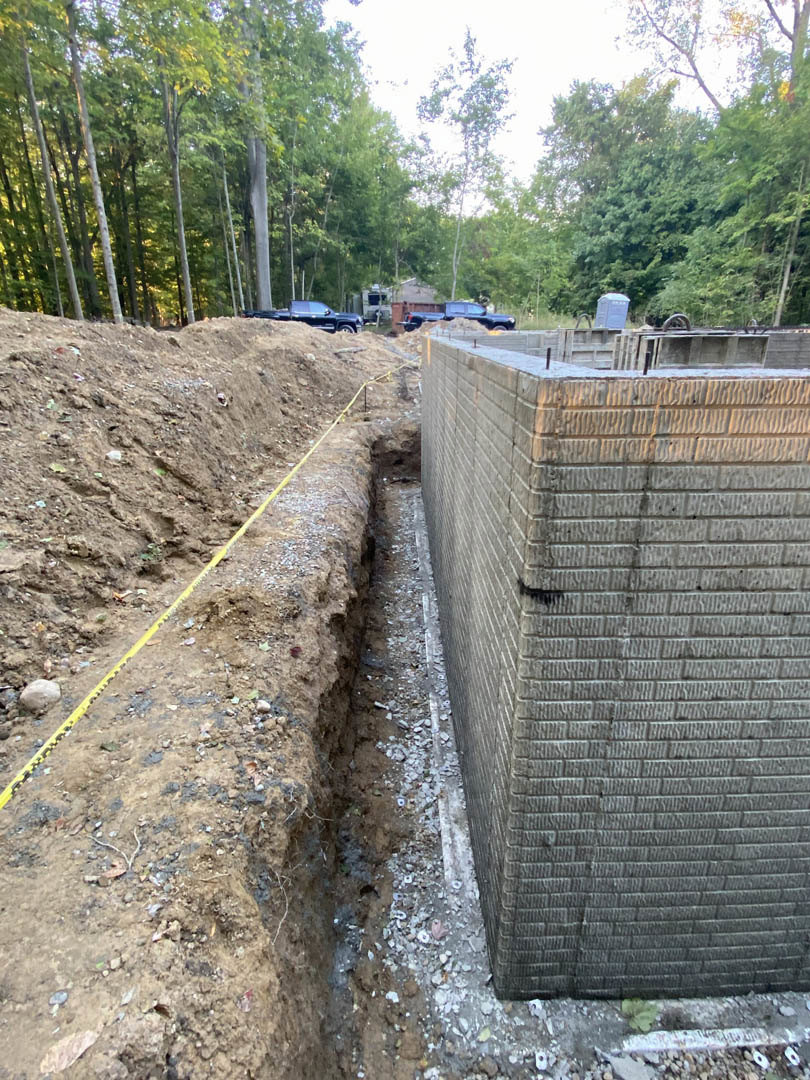 Concrete block wall beside a trench with yellow caution tape, surrounded by parked cars and trees in a forested outdoor setting