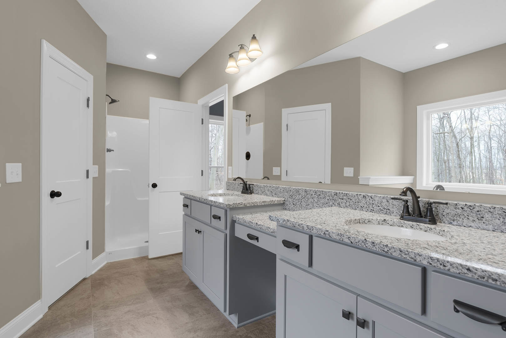 Bathroom with granite countertops, dual sinks, white cabinetry, tile flooring, three-light fixture above mirror, window overlooking trees, white door with silver handle, white wall