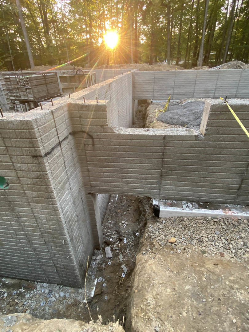 Framed custom home under construction with exposed beams, stacked lumber, rocky ground, and sunlight filtering through surrounding trees