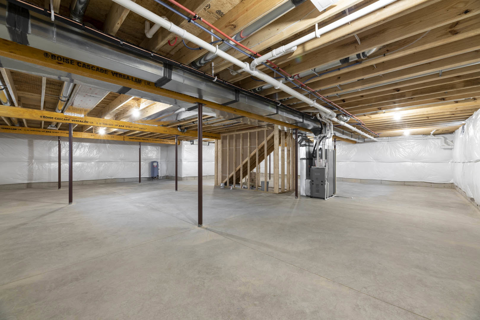 Basement with exposed pipes, wooden ceiling beams, concrete floor, and framed staircase; close-up of a grey utility box.