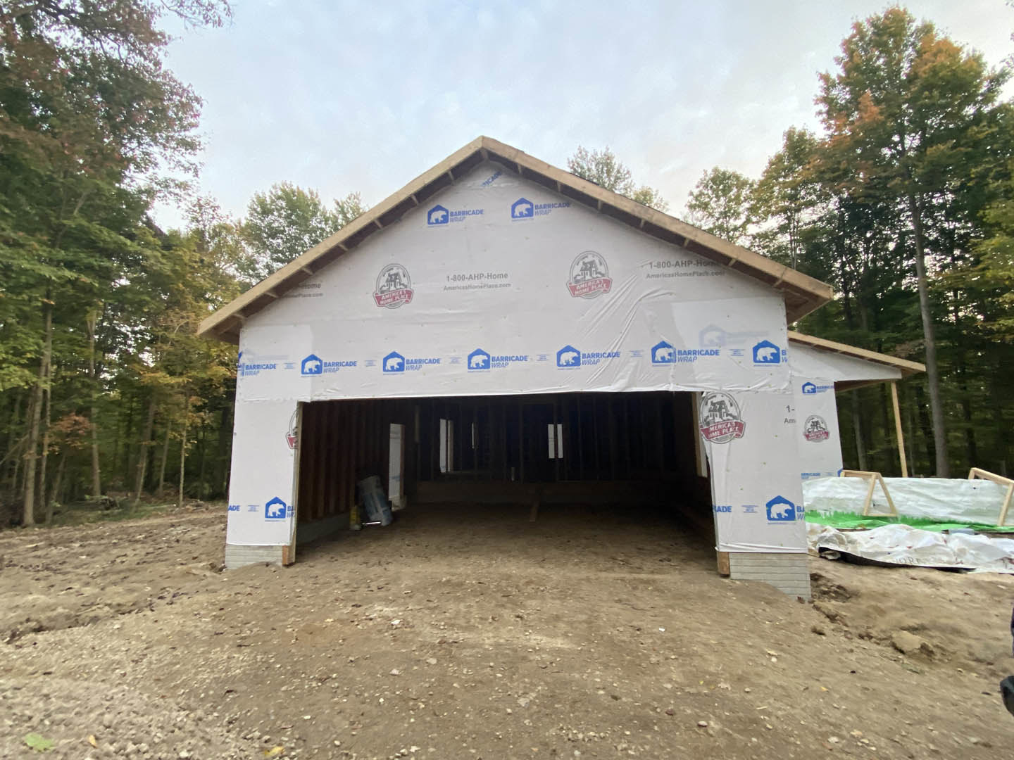 Framed custom home under construction with exposed wood beams, white weatherproof sheeting, dirt floor, and surrounding trees in the background