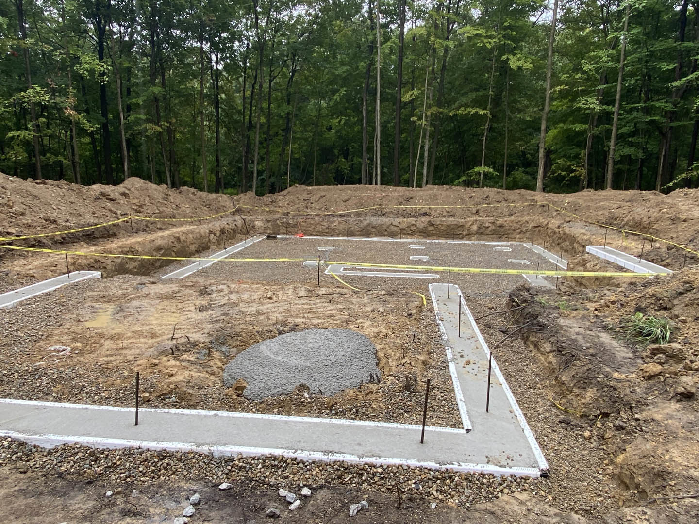 Dirt construction site with poured concrete footers, yellow caution tape, and forested trees in the background