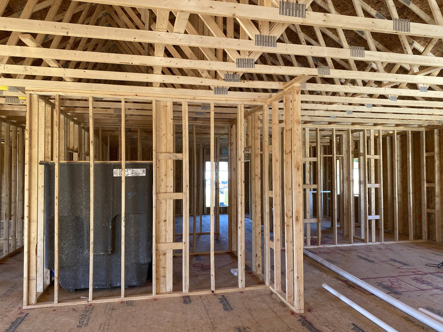 Exposed wood framing and beams with metal studs, unfinished wooden roof structure, white plumbing pipes laid across subfloor, grey metal wall panel with white label, partially