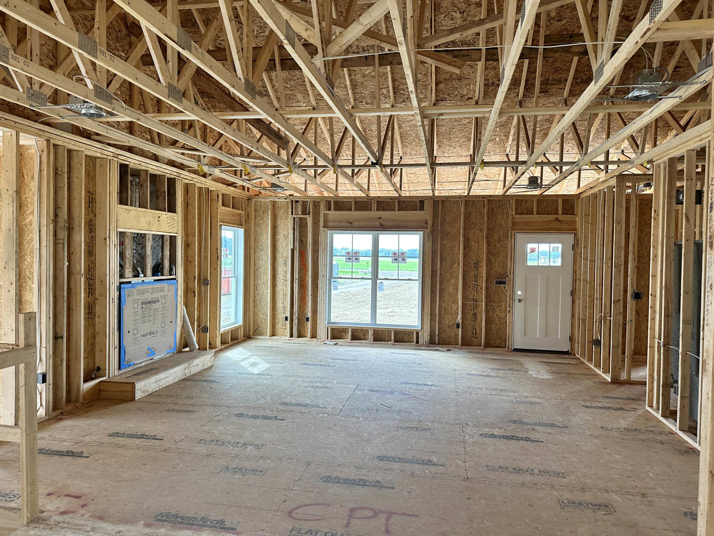 Unfinished room with exposed wooden ceiling beams, white door with glass window, large window with taped cardboard, wood paneling, and construction materials on the floor