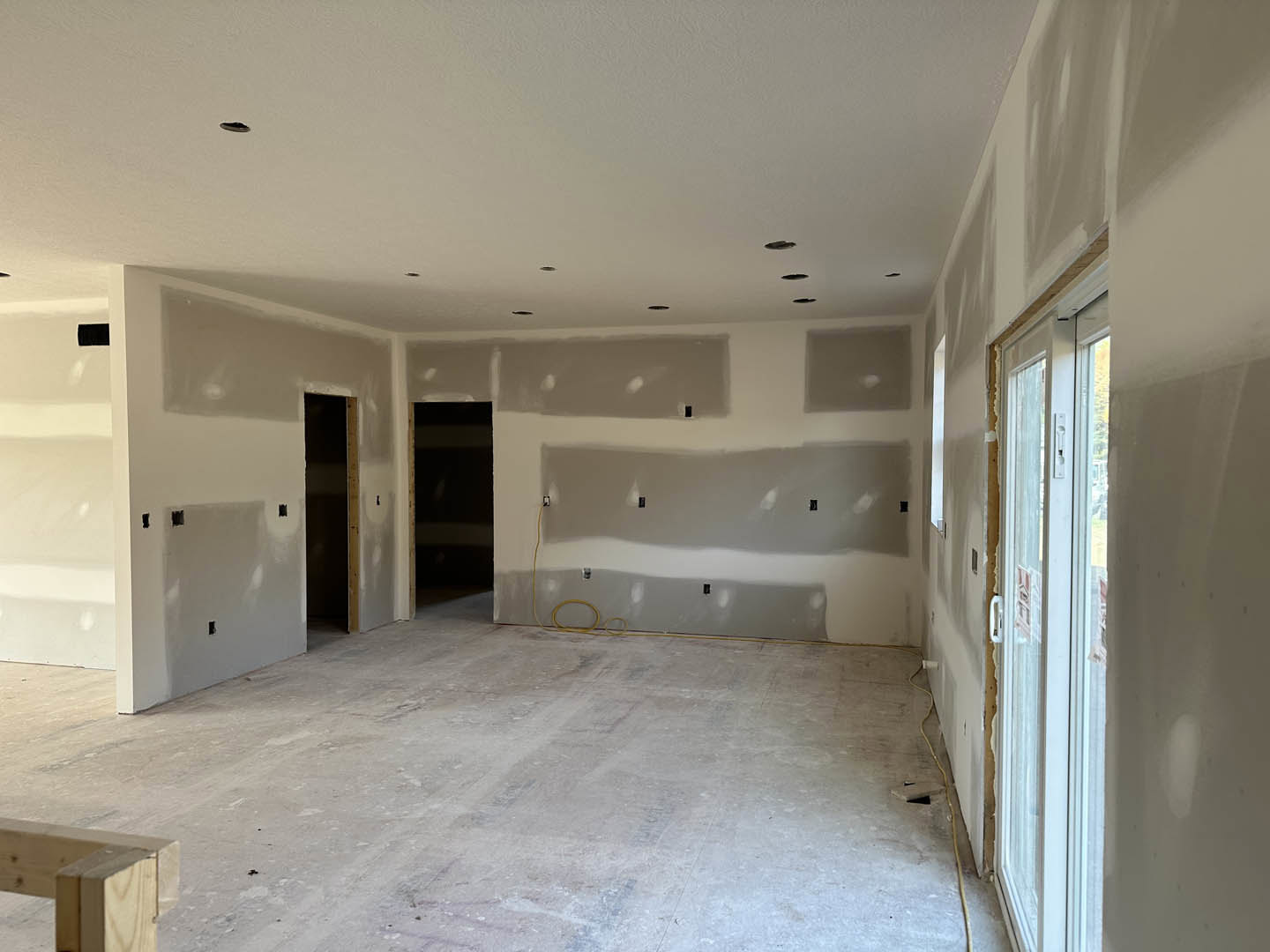 White plaster walls, black door with wooden frame, concrete floor, yellow hose, black tape on wall, empty room.
