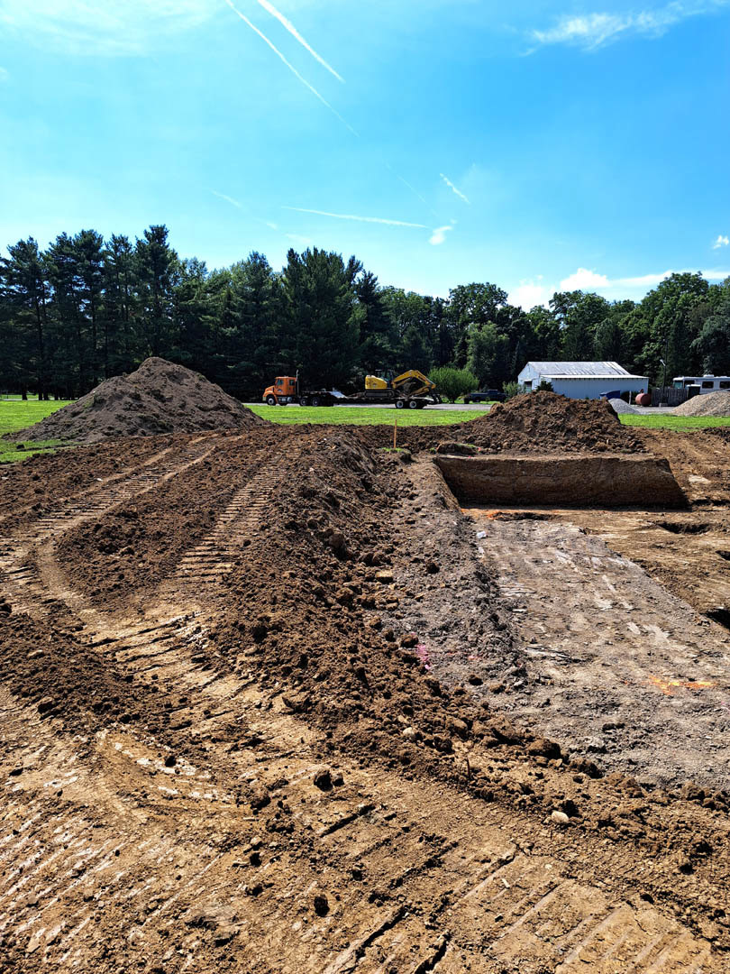 Dirt piles and tire tracks on a construction site with a tractor, white building with blue roof in the background, blue sky, scattered clouds, and surrounding trees
