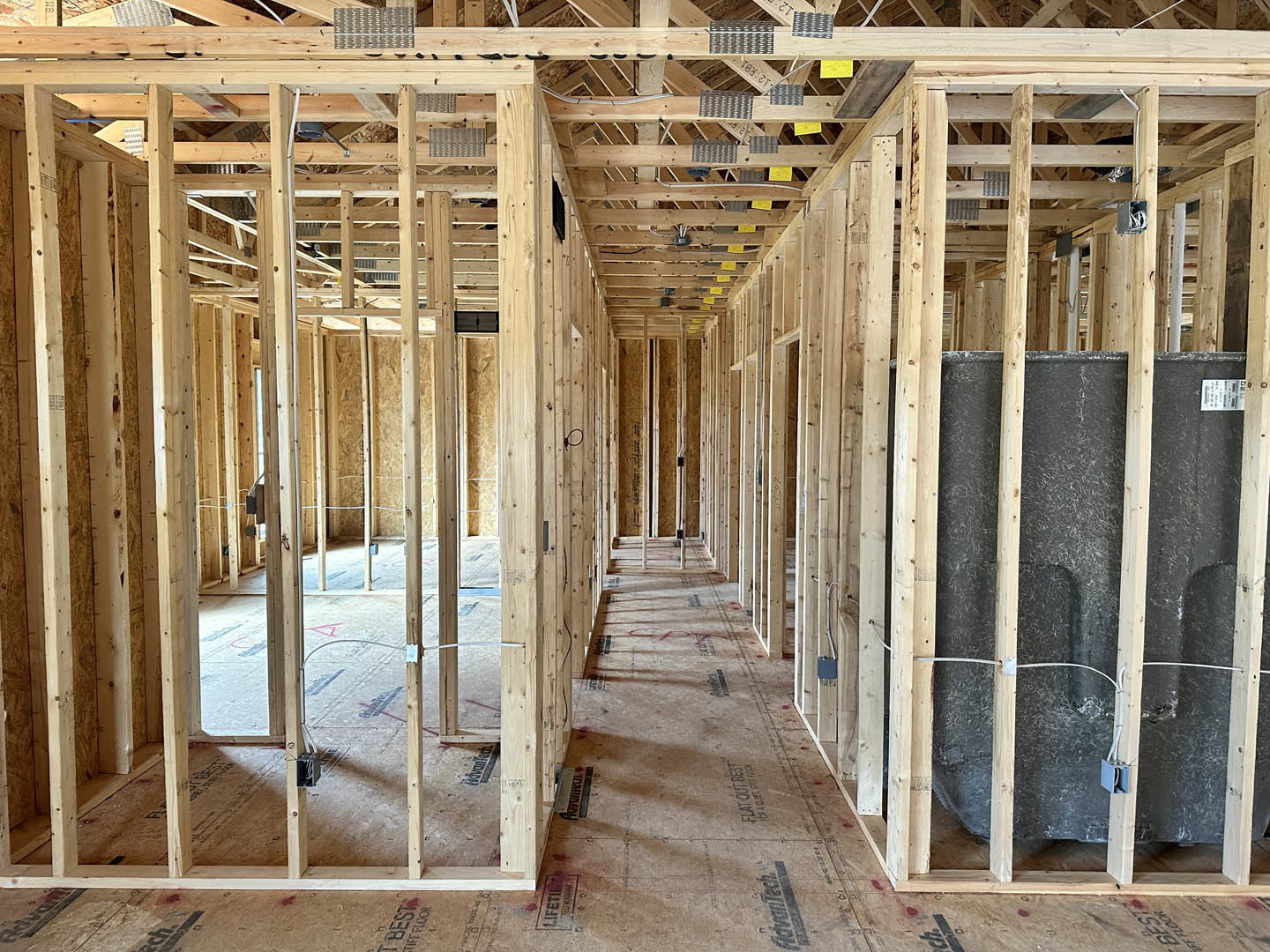 Wood-framed interior wall with exposed beams, black pegs, stacked boxes, insulation, and metal pipes under unfinished roof structure