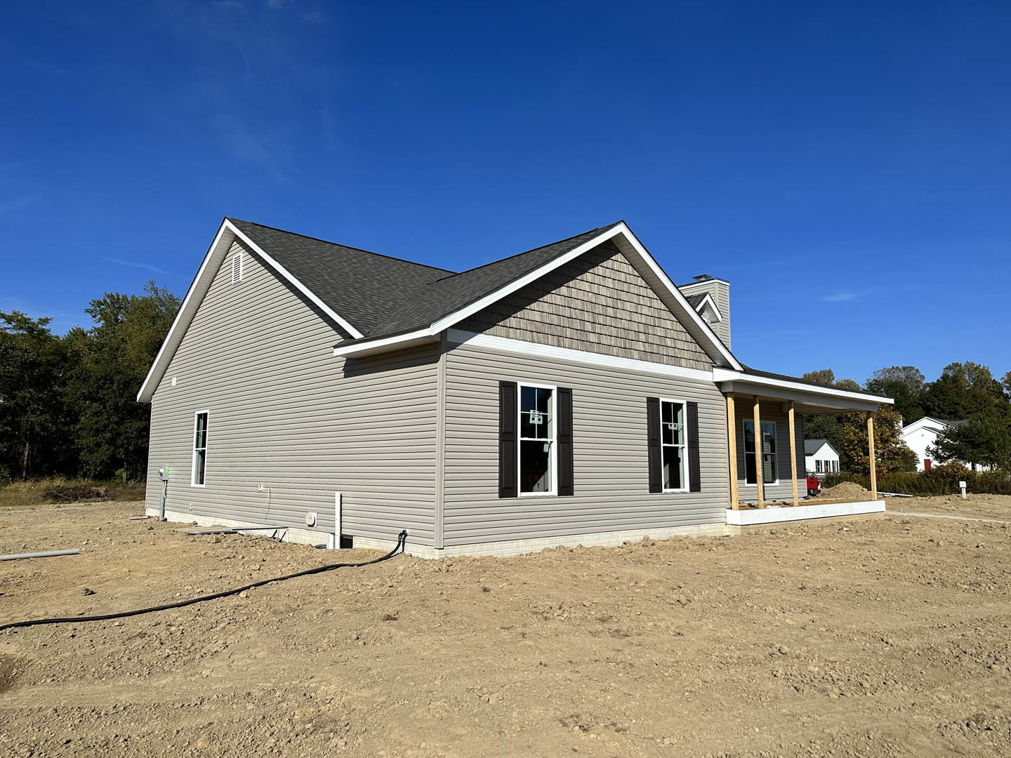 Partially built house with exposed framing and white window trim, surrounded by dirt and construction materials, hose emerging from ground, blue sky overhead