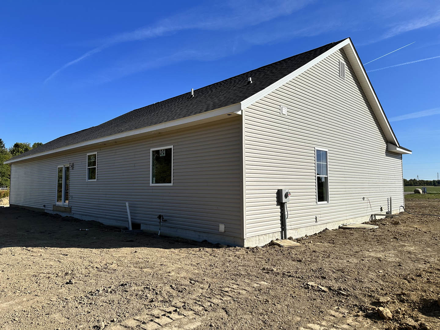 Wood-framed house under construction with exposed sheathing, dirt lot in foreground, blue sky overhead, and historic Rockingham Meeting House visible in the background