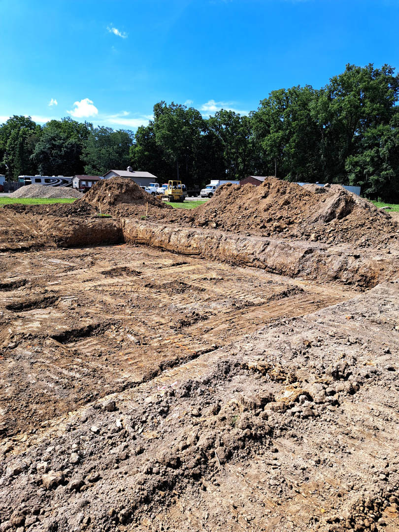 Bulldozer parked beside a large pile of dirt on a construction site, square excavation in soil, trees and blue sky with clouds in background