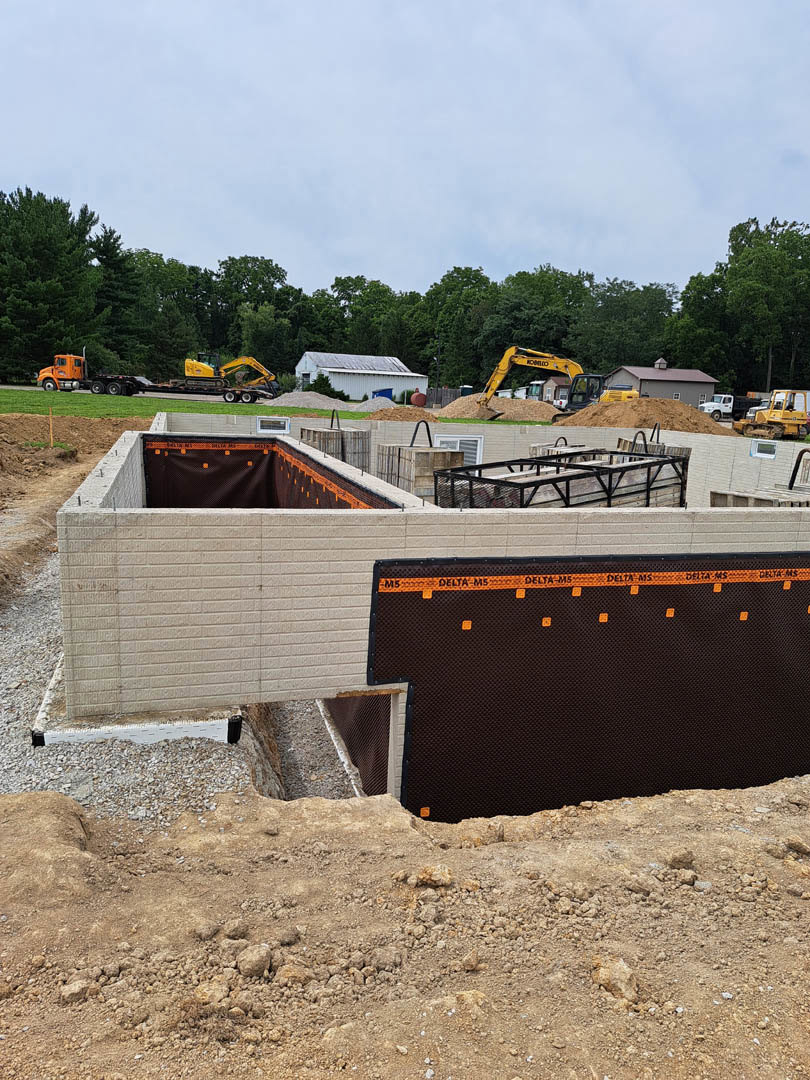 Concrete foundation surrounded by exposed soil, construction materials, and a blue shipping container; white building partially visible, trees and clear sky in background.