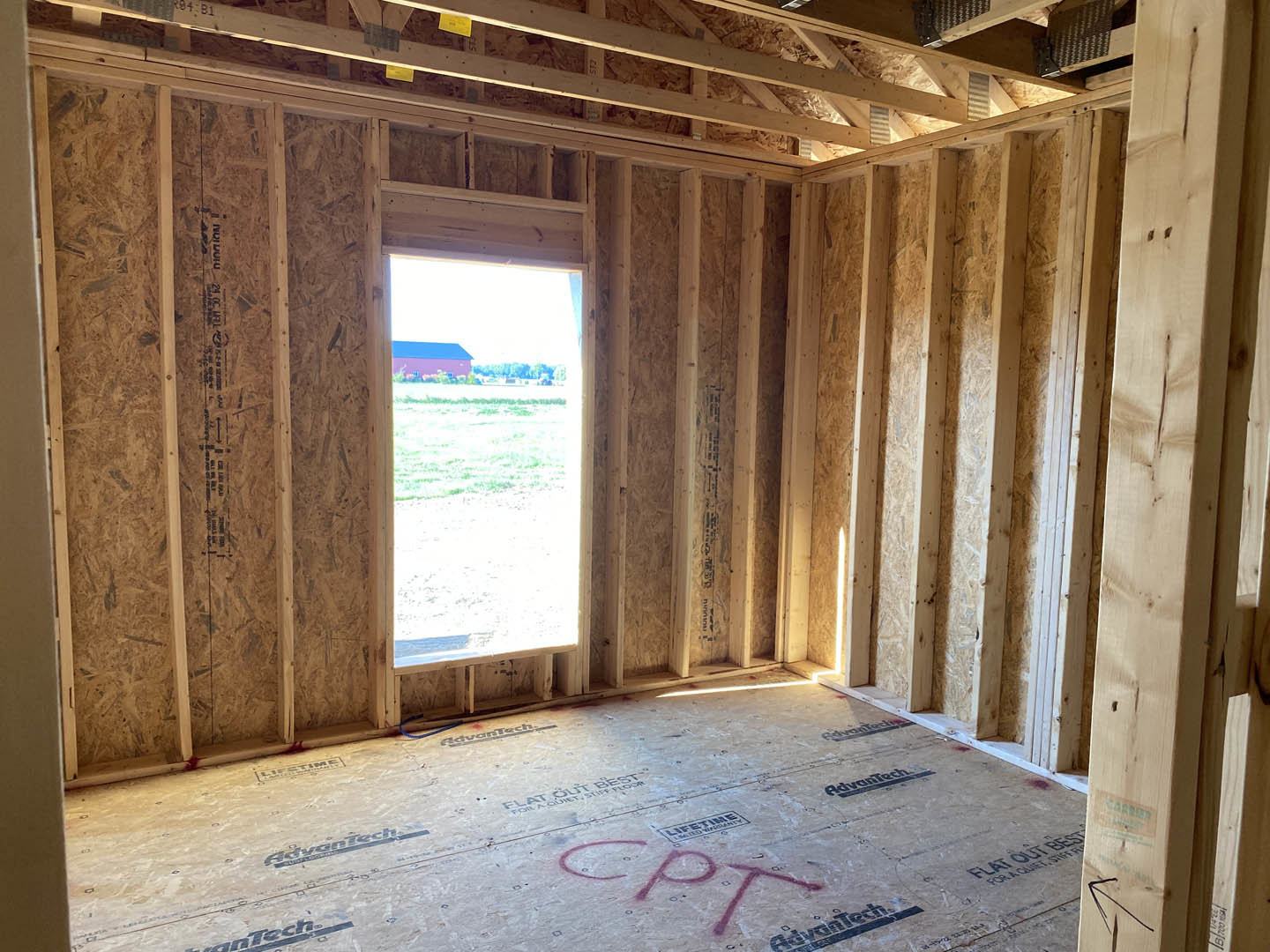 White-walled room with hardwood flooring, single window showing red building outside, and wooden door with brushed metal handle