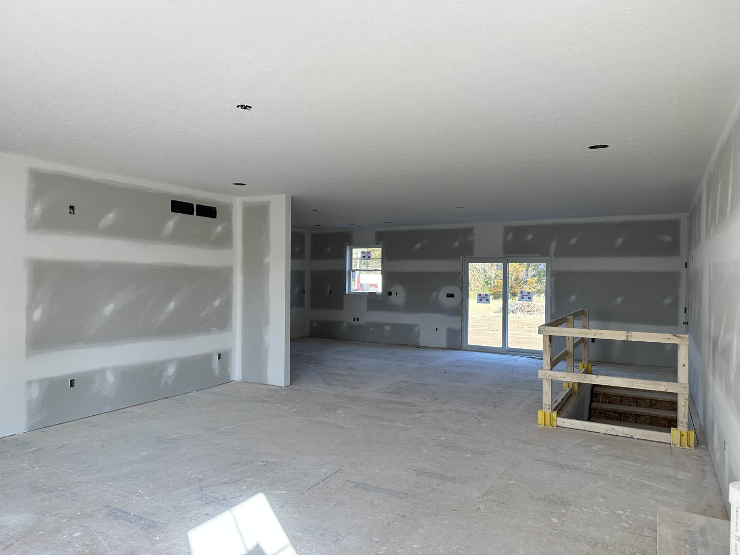 Open room with white and gray plaster walls, concrete floor, wood ladder leaning against wall, large windows and glass door showing tree outside