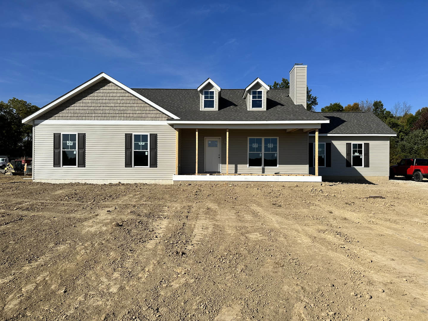 Two-story house under construction with exposed siding, white double front doors, covered porch, dirt yard, and red pickup truck parked nearby.