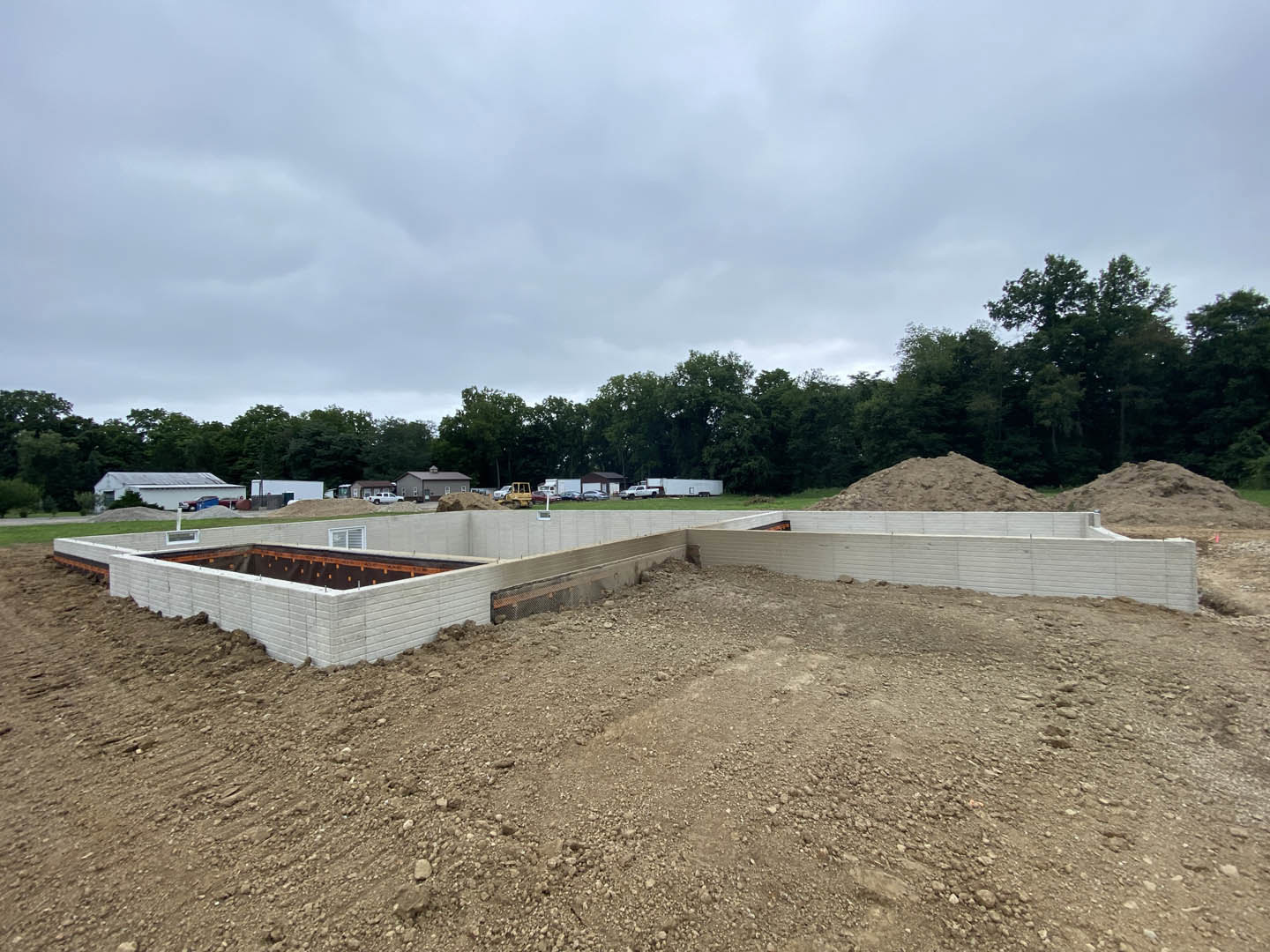 Concrete foundation surrounded by dirt and gravel, construction materials scattered, mature trees and cloudy sky in background