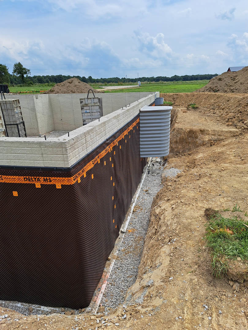 Large concrete foundation surrounded by dirt and gravel, black mesh covering part of a wall, metal barrel in foreground, patch of grass near fence, blue sky with scattered clouds