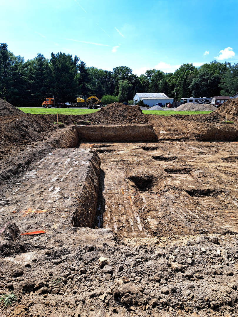 White custom home with blue roof surrounded by dirt construction site, mature trees in background, clear blue sky overhead
