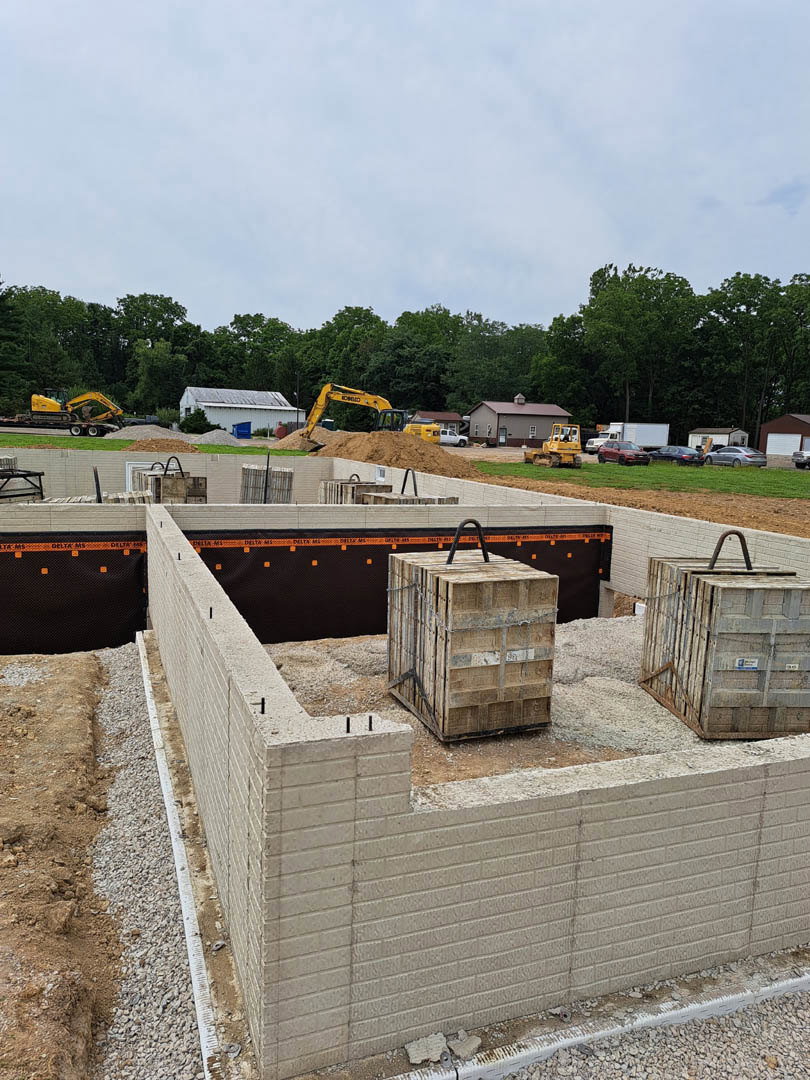 Concrete foundation surrounded by stacked wooden crates, white building with blue bin, black and orange construction barrier, blue sky with scattered clouds