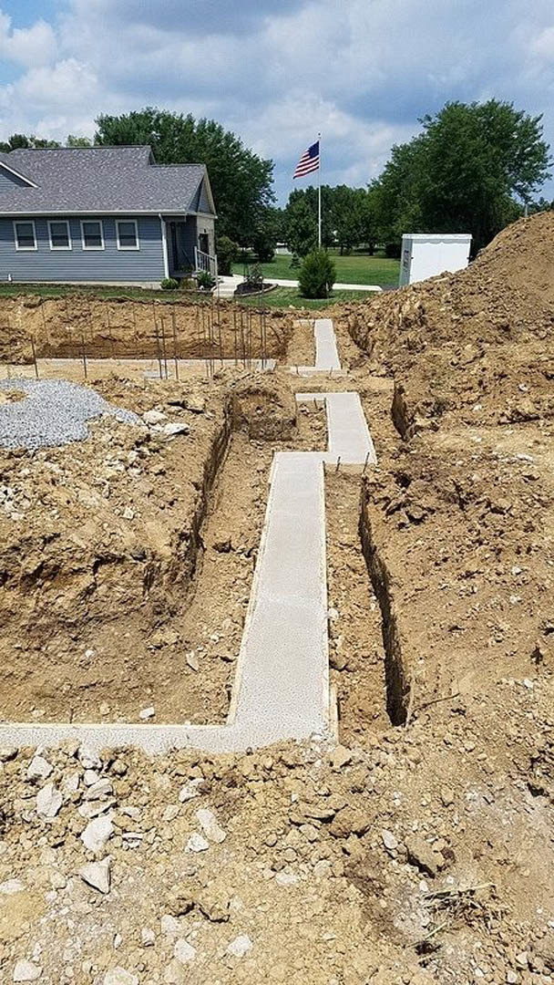 Concrete walkway surrounded by bare soil, white house with few windows, white exterior wall, large tree, rocky landscaping, and distant hill under partly cloudy sky