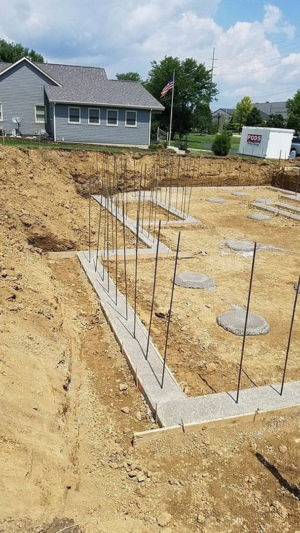 Concrete foundation under construction with exposed steel rebar, adjacent to a partially built house with several windows, white tent featuring a red sign, flagpole, and scattered