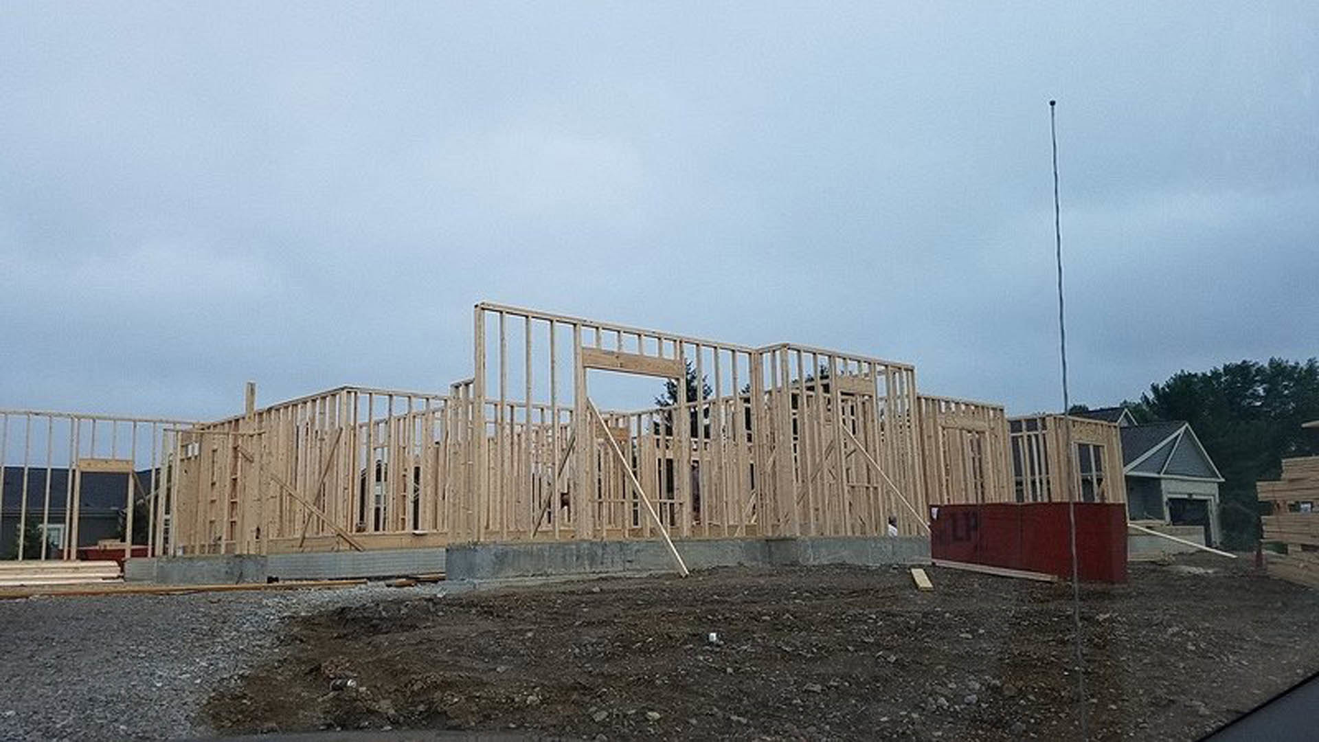 Wooden framing and metal scaffolding surround a partially constructed house, red gate with white trim in foreground, cloudy sky overhead.