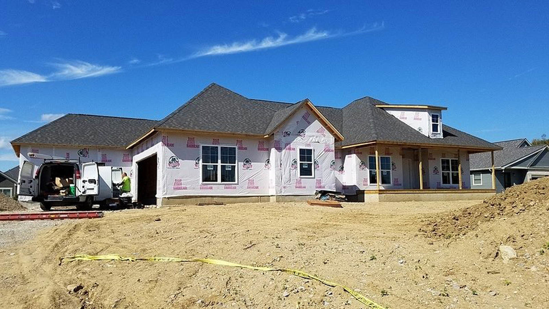 Partially built house with exposed framing, unfinished exterior walls, and a man in work clothes standing near the open rear door of a van on a dirt lot under a cloudy sky