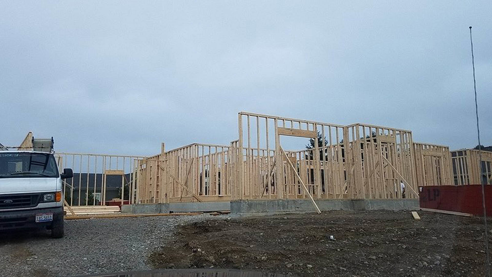 Wood-framed structure under construction with exposed beams, parked white van in foreground, cloudy sky overhead