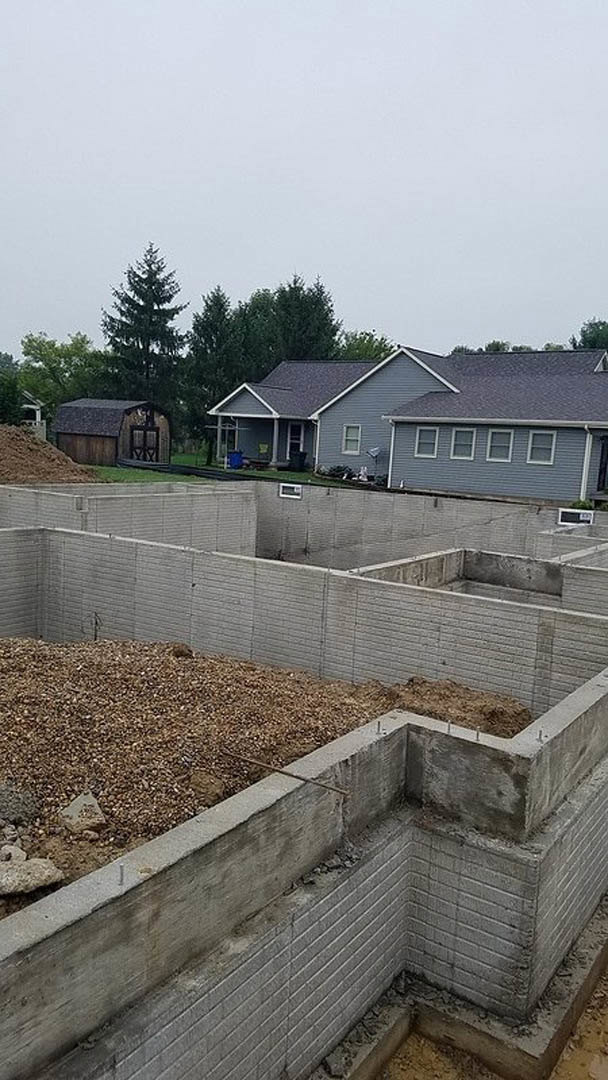 Grey-roofed house with small wooden barn, open door, concrete foundation, piles of dirt and gravel, close-up rock, and concrete wall with ground hole; neighboring houses and trees