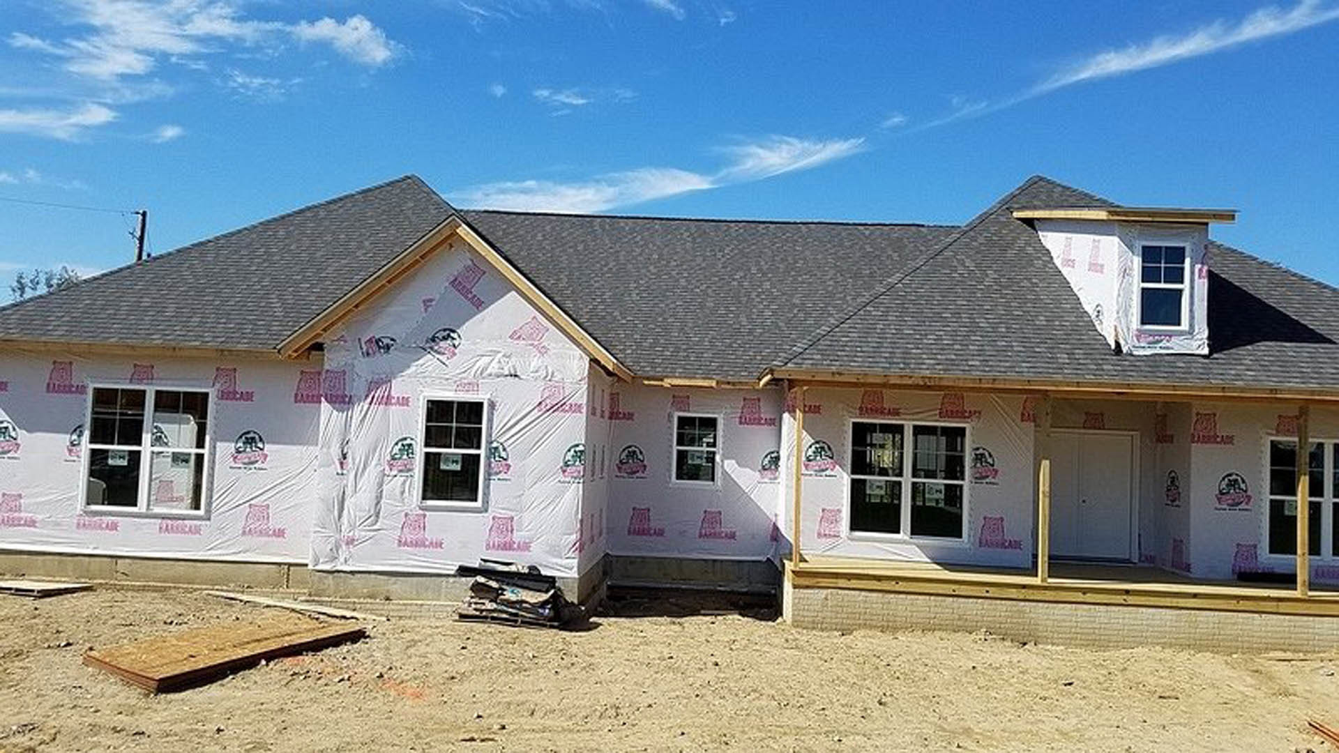 Two-story home under construction with plastic sheeting covering windows and entry, exposed framing, and unfinished exterior walls under a cloudy sky