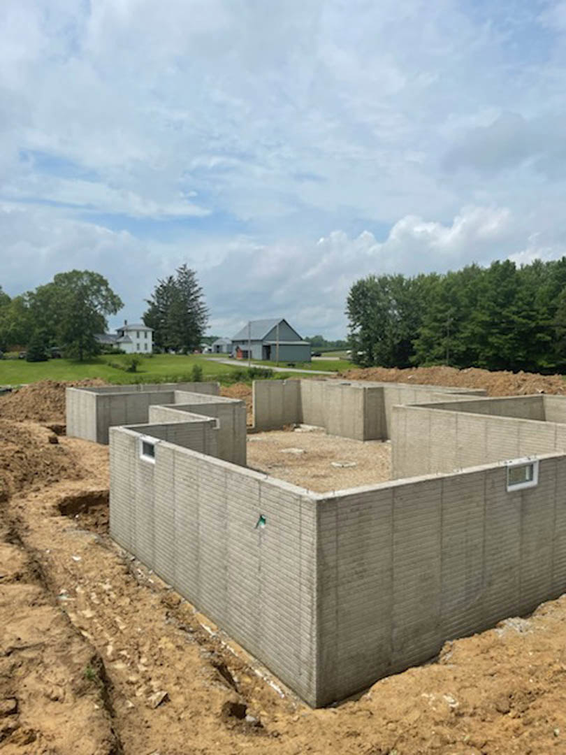 Concrete foundation with block walls set in a grassy field, surrounded by trees under a cloudy sky, with a grey barn visible in the background.