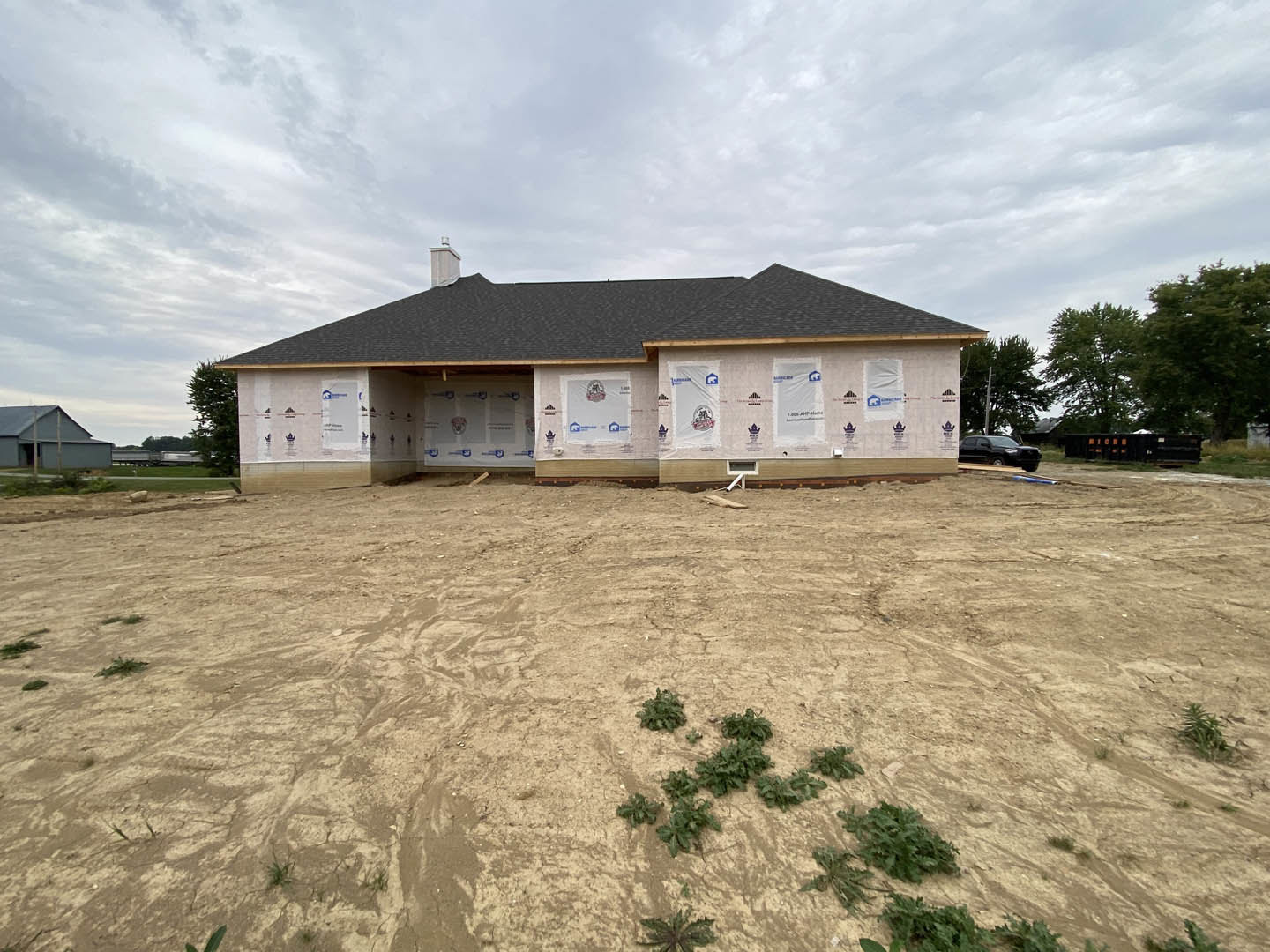 Partially built house with exposed framing, plastic coverings, dirt lot, scattered construction materials, surrounding trees, and overcast sky