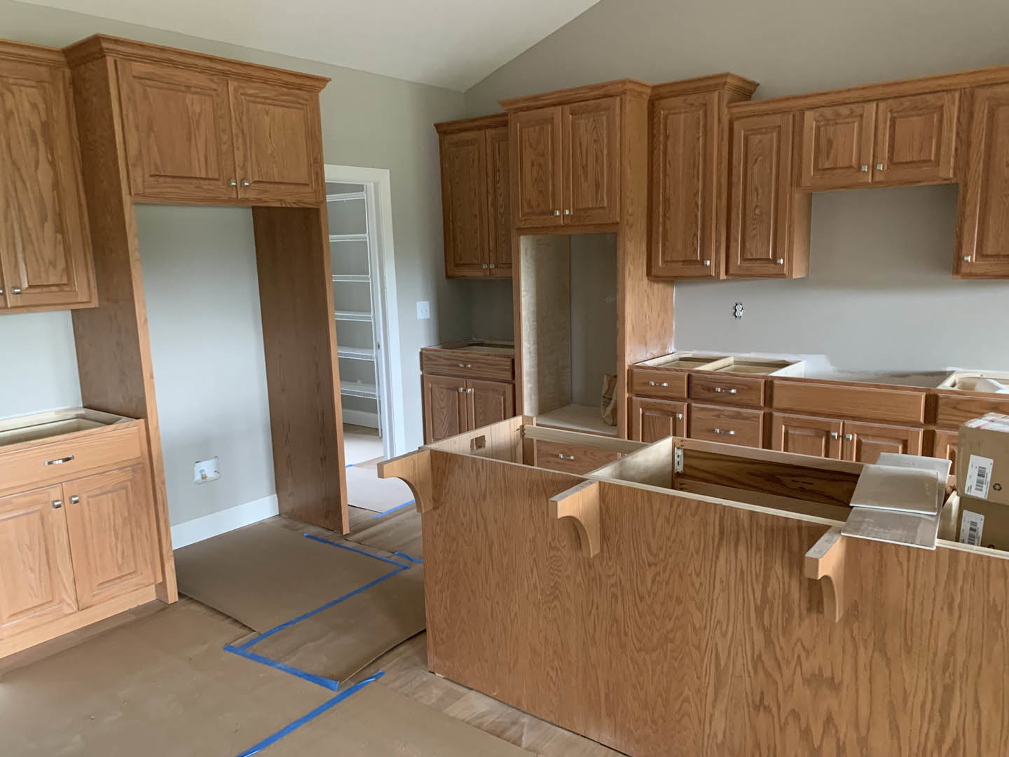 Kitchen with natural wood cabinets, white walls and trim, light-colored countertop, door leading to adjacent room, cardboard sheet on the floor