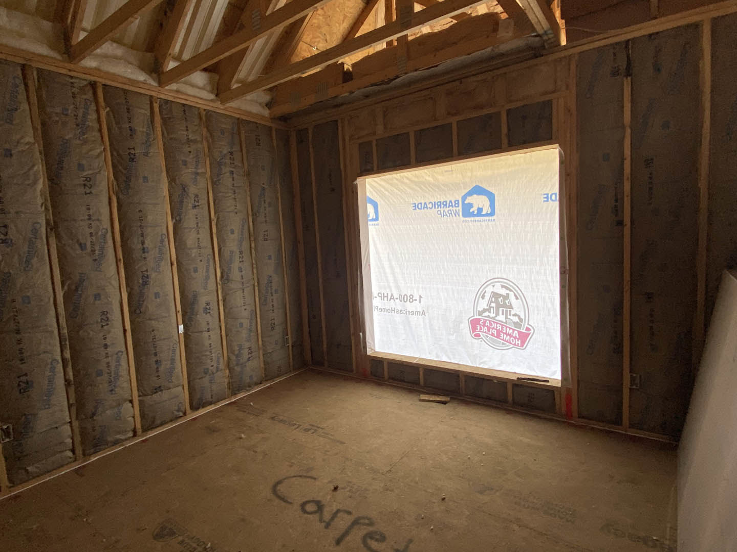 Unfinished room with exposed ceiling beams, white insulated walls, and a window covered in protective plastic; handwritten text and blue logo visible on floor and wall.