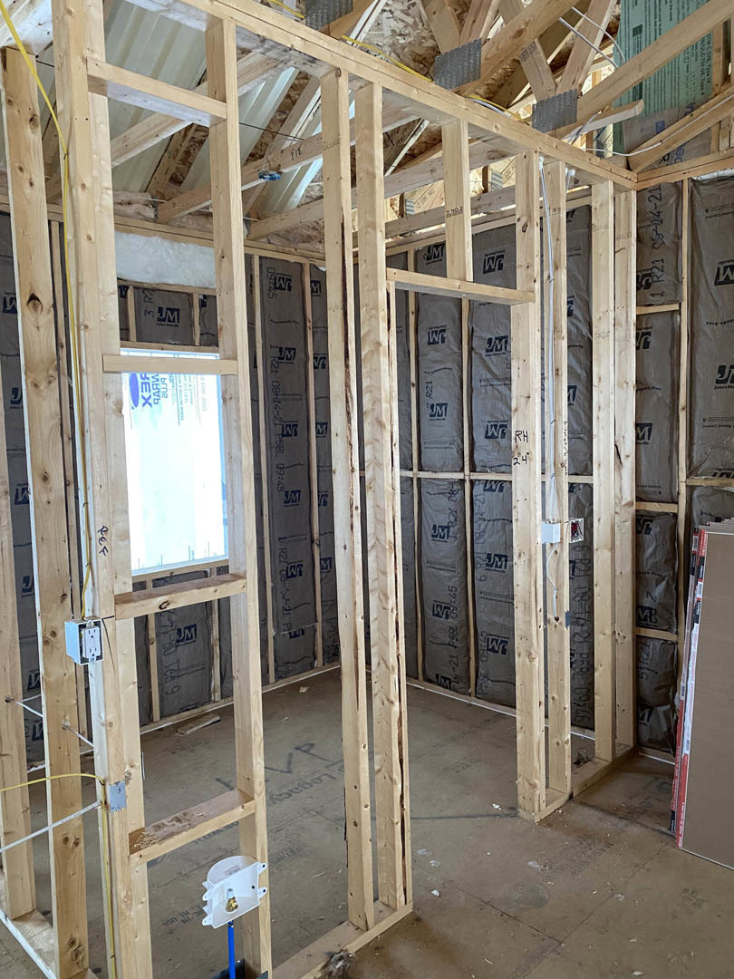 Wood-framed room under construction with exposed beams, insulation, a window, a white electrical outlet with a plug, and a person standing near the window.