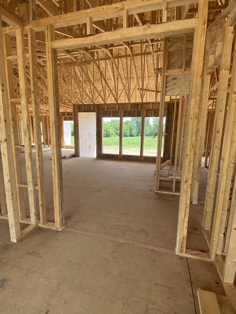 Exposed wood framing and beams inside a partially constructed home, with a window overlooking trees and a white insulated surface marked with black and red lines.