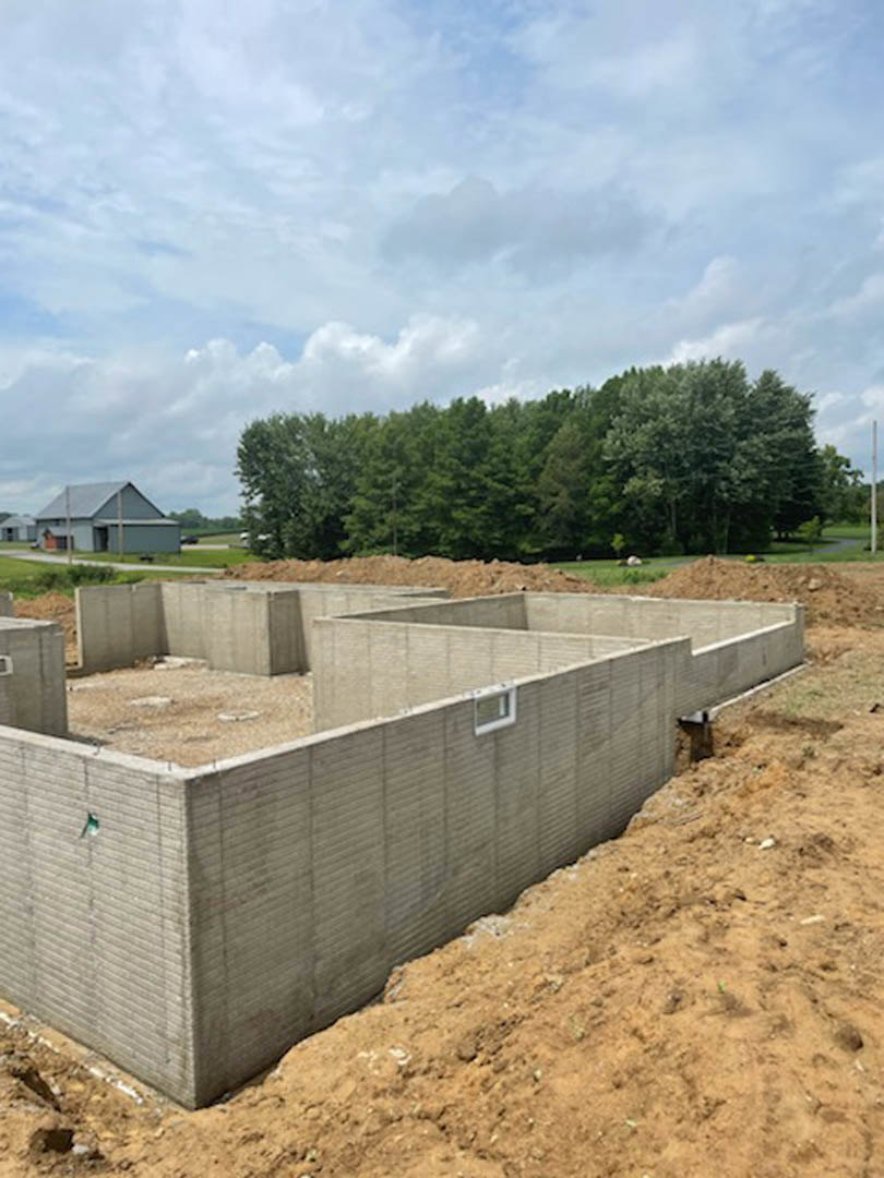 Concrete foundation slab set in a dirt field, bordered by scattered trees and under a partly cloudy blue sky
