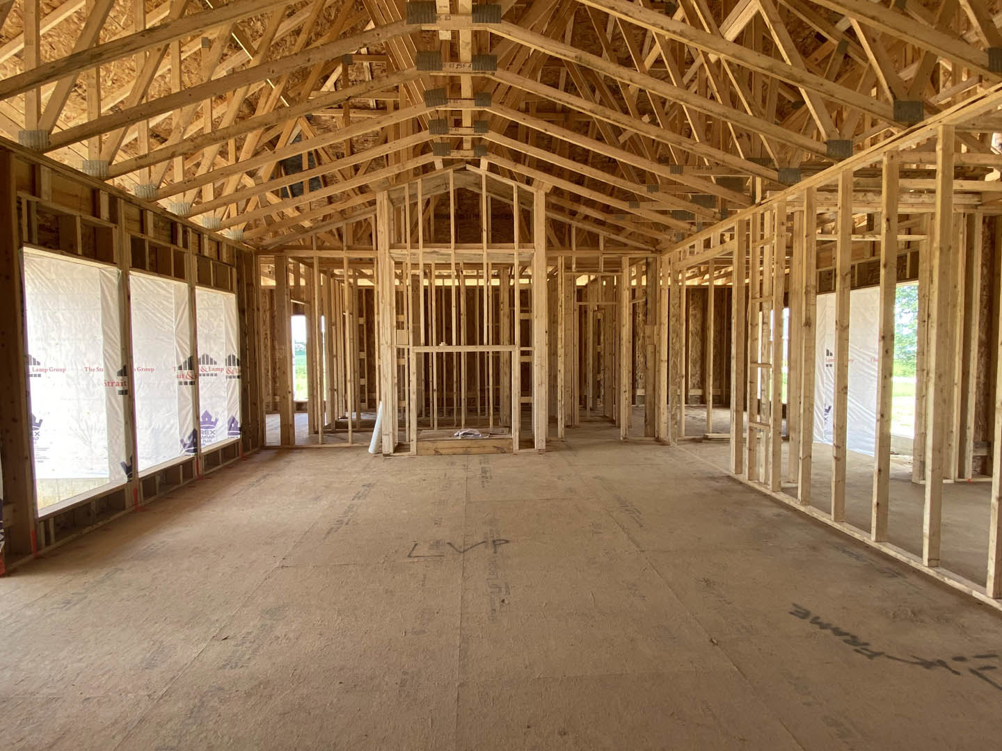 Open room with exposed wooden ceiling beams, large window letting in daylight, light wood flooring, white walls, and visible wood framing