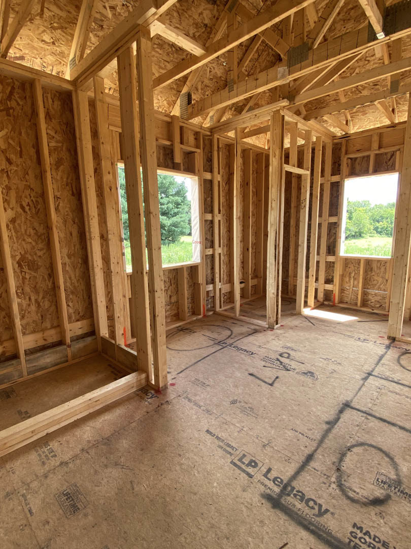 Exposed wood framing in an unfinished room, large window with view of trees, construction materials and bag on plywood floor