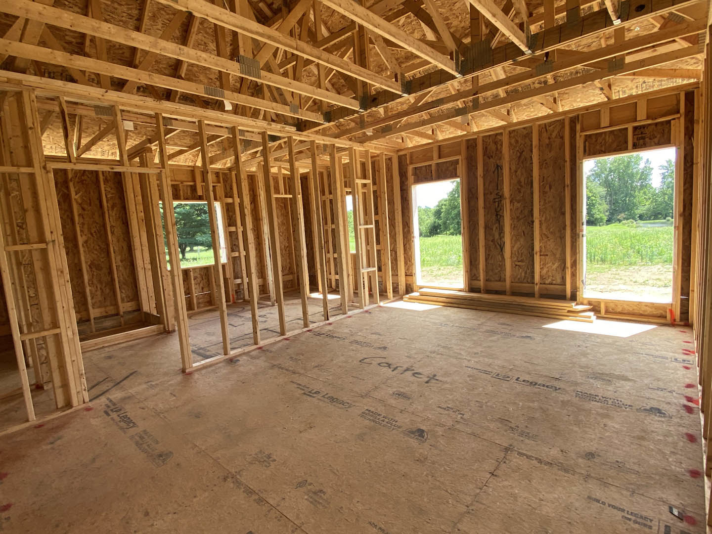 Living room with exposed wood ceiling beams, large windows overlooking grassy field and forest, hardwood floors, and partial view of staircase.