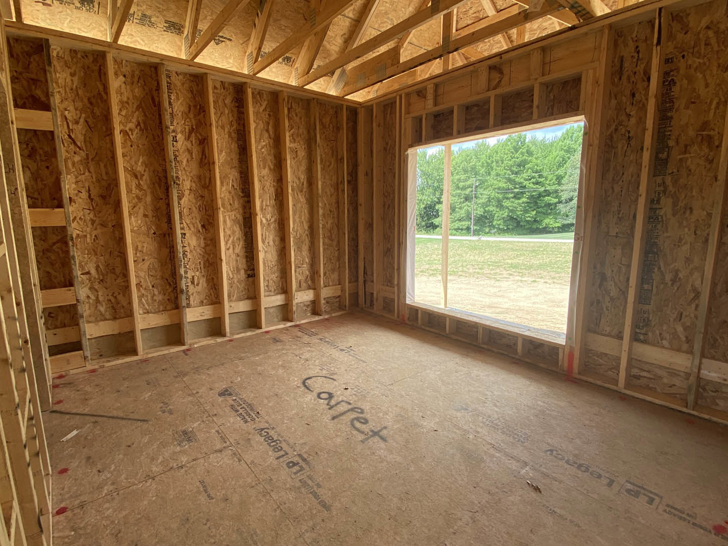 Room under construction with exposed wood framing, large window overlooking trees, unfinished wood floor