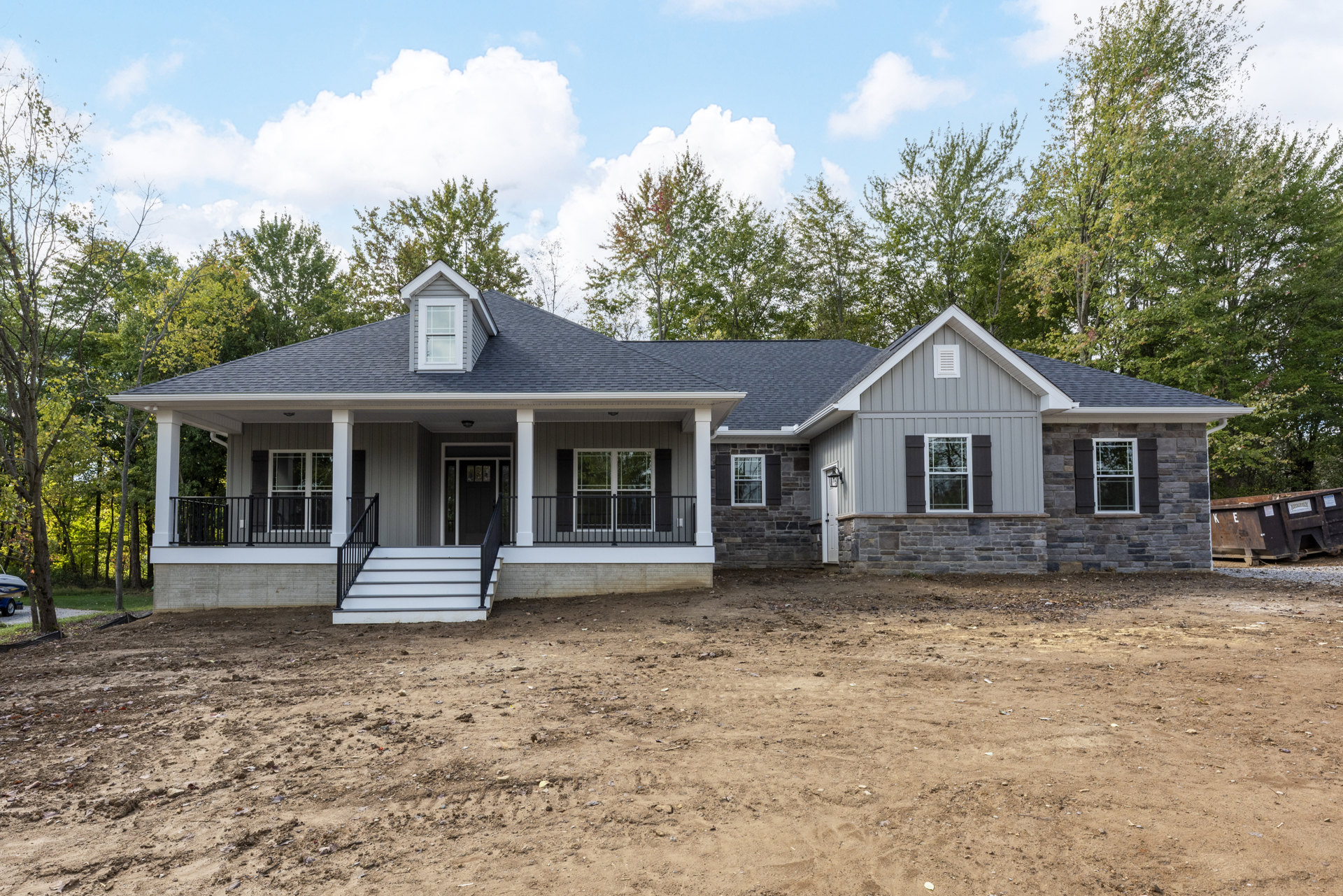 Modern house exterior with white-framed windows, black metal railings, and a dirt yard in front; cloudy sky and trees in background