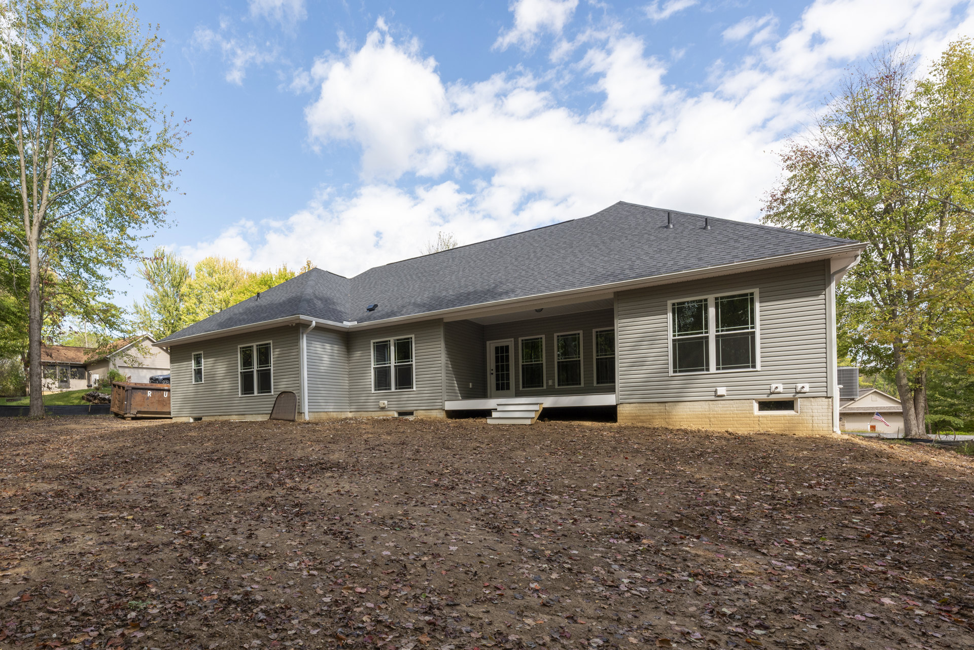 Two-story home with white-framed windows, covered porch, metal gate, and bare dirt backyard under a partly cloudy blue sky; scattered leaves near the foundation.