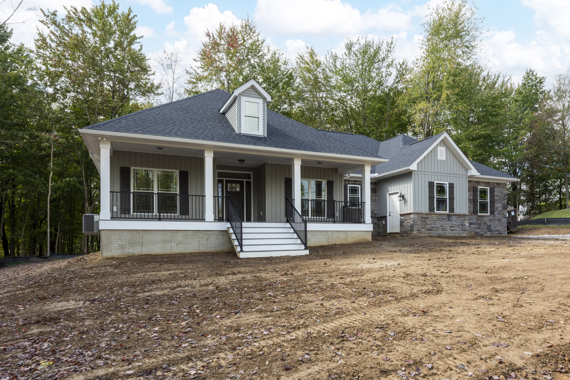 Two-story house with white siding, black glass-paneled front door, covered porch with stairs, dirt yard in foreground, trees and small white house in background, roof window