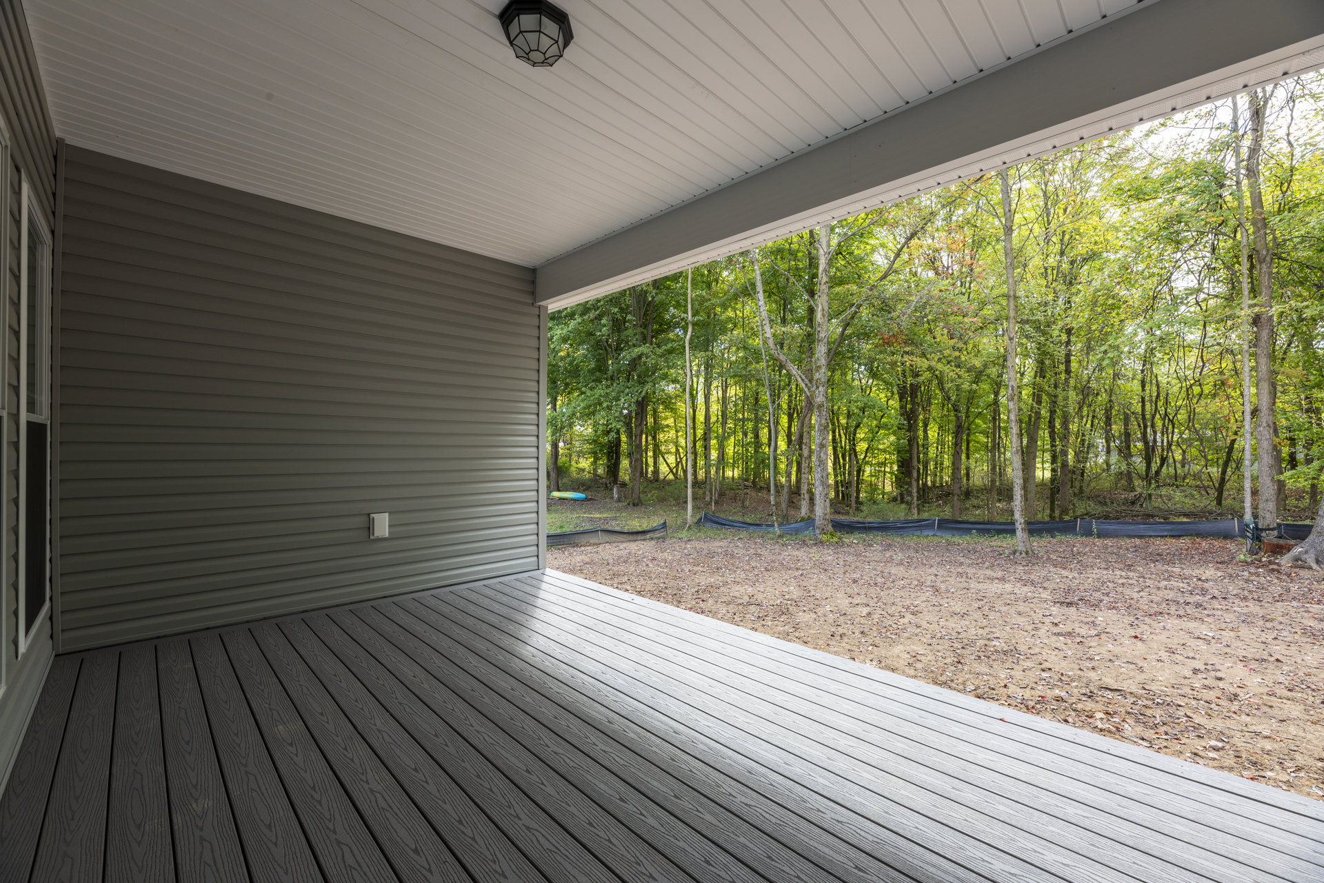 Wooden deck with composite railing overlooking trees and bare ground, shaded by ceiling, outdoor chair near tree, blue trampoline visible in wooded area.