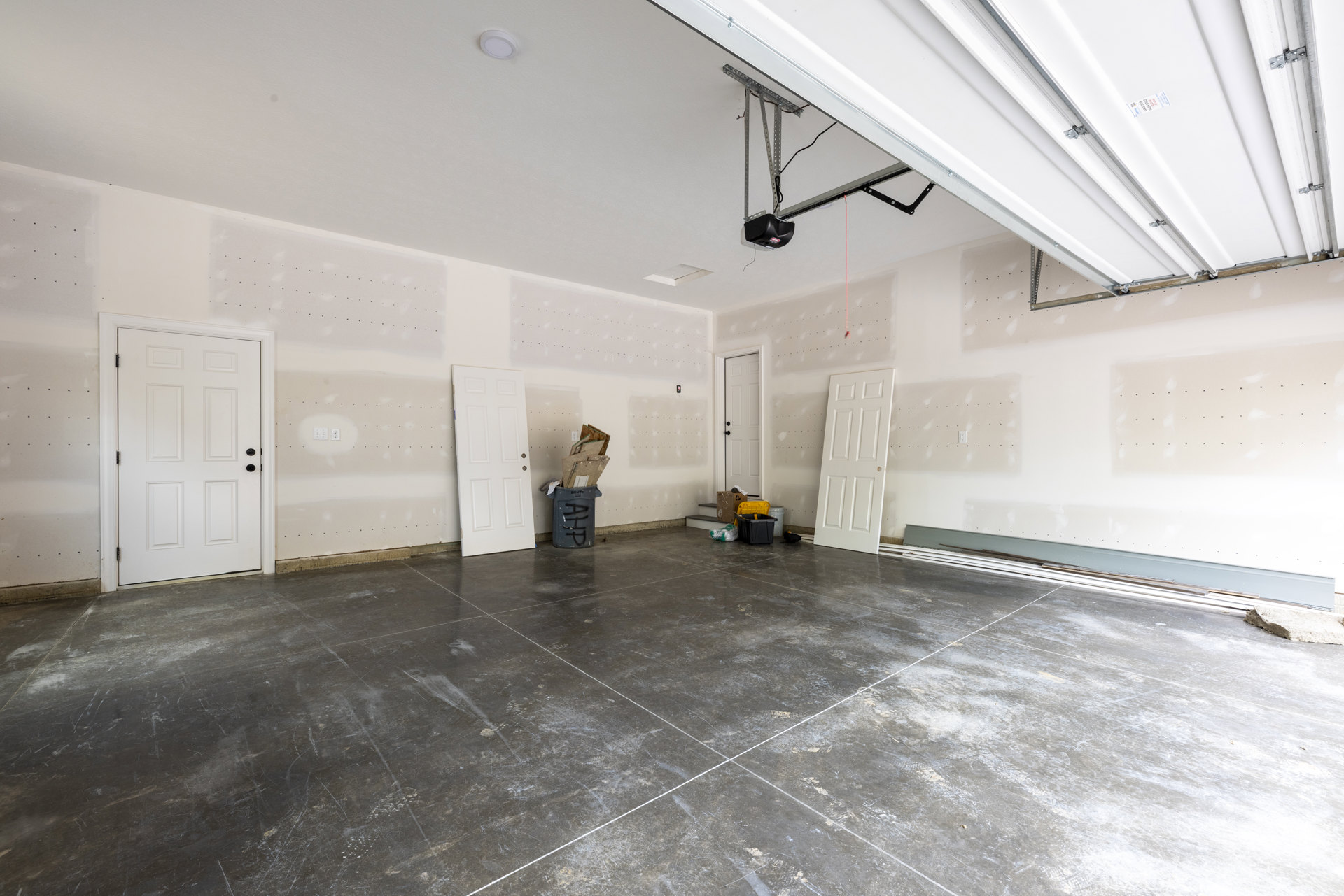 White walls and ceiling, white door with black knobs and three holes, grey tile floor with white grout lines, garage door visible in background.